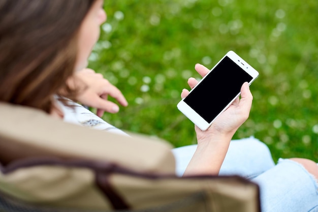 Giovane donna freelance seduta su una sedia e che utilizza lo smartphone Lavoro a distanza e relax nella natura Sfondo dello schermo nero per il testo