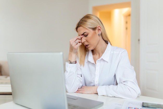 Giovane donna frustrata che lavora alla scrivania dell'ufficio davanti al computer portatile che soffre di mal di testa cronico quotidiano trattamento online nominando a una consulenza medica radiazioni elettromagnetiche paga per malattia