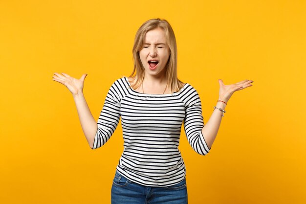 Giovane donna frustrata in abiti a righe che urla tenendo gli occhi chiusi, allargando le mani isolate su sfondo giallo arancione in studio. Persone sincere emozioni, concetto di stile di vita. Mock up spazio di copia.