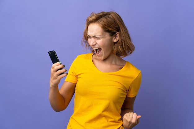 Giovane donna georgiana isolata su sfondo viola usando il telefono cellulare e facendo il gesto della vittoria