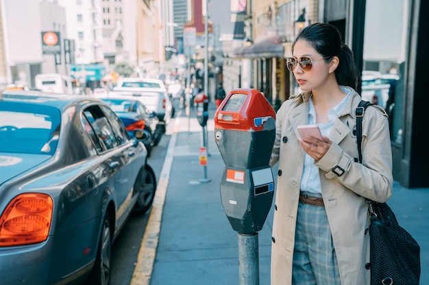 Giovane donna giapponese asiatica affascinante che paga per il parcheggio dell'auto alla macchina con l'app online del telefono cellulare. bellissimo stile di vita locale della signora dell'ufficio a san francisco negli stati uniti. cellulare femminile della holding che effettua il pagamento