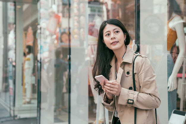 Giovane donna giapponese asiatica che utilizza smartphone mentre si trova nel quartiere dello shopping della città con una vetrina di un negozio di moda sullo sfondo. ragazza sorridente che cerca la direzione sul telefono cellulare nell'urbano moderno.