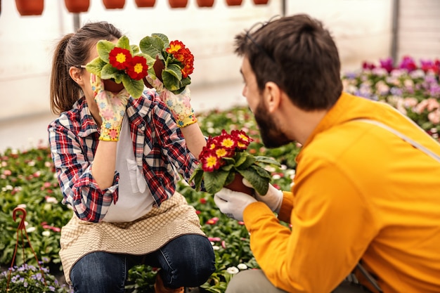 Giovane donna giocosa accovacciata e tenendo vasi con fiori e scherzare. Intrattiene il suo collega.