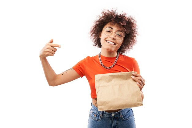 Giovane donna gioiosa con riccioli afro in vestiti arancioni che tengono pacchetto artigianale isolato su sfondo bianco studio, consegna cibo, borsa ecologica, ragazza caucasica kazaka con regalo