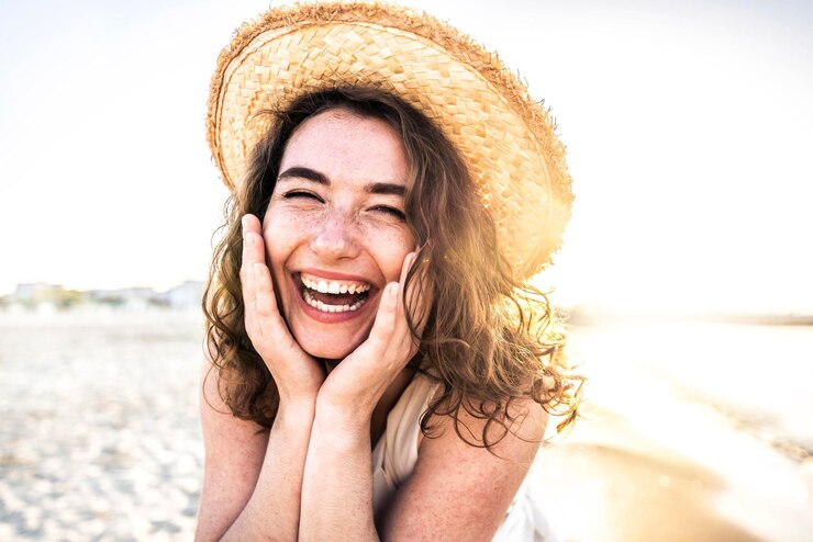 Giovane donna gioiosa in camicia bianca che indossa il cappello che sorride alla macchina fotografica sulla spiaggia