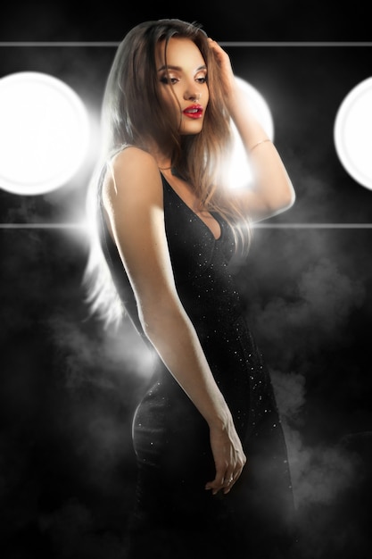 Giovane donna glamour con le labbra rosse in abito stretto nero che osserva da parte in studio sulla parete scura con luci e fumo