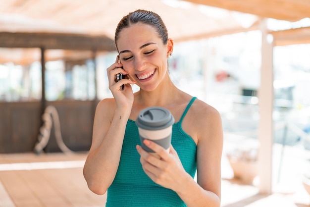 Giovane donna graziosa all'aperto utilizzando il telefono cellulare e tenendo un caffè con espressione felice