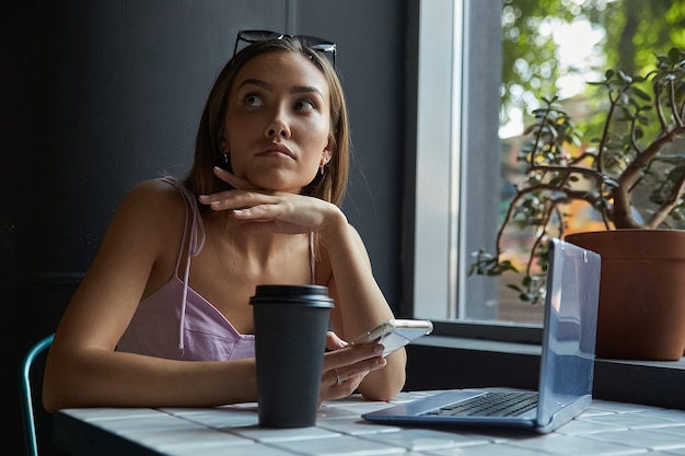 Giovane donna graziosa asiatica che si siede alla tavola del caffè con il computer portatile facendo uso dello smartphone