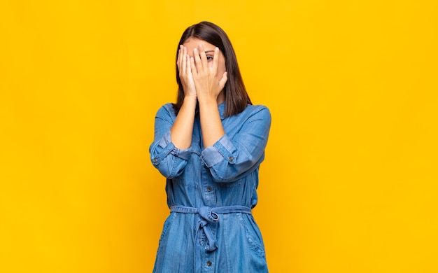 Giovane donna graziosa che copre il viso con le mani, sbirciando tra le dita con espressione sorpresa e guardando al lato