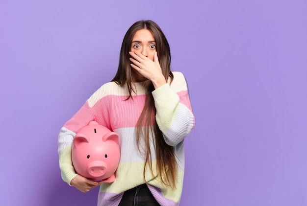 Giovane donna graziosa che copre la bocca con le mani con un'espressione scioccata e sorpresa, mantenendo un segreto o dicendo oops