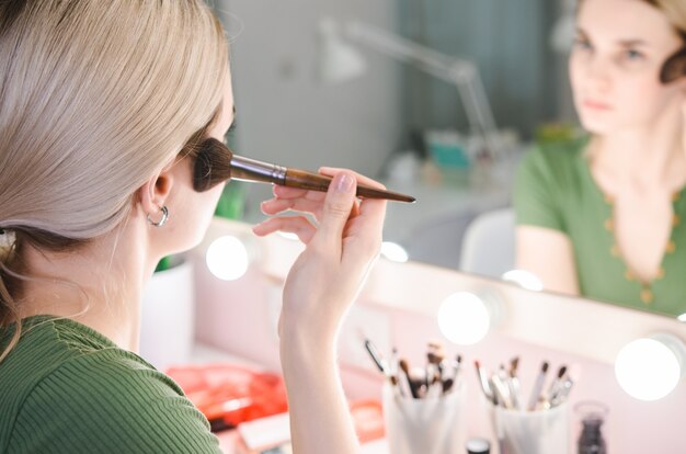 Giovane donna graziosa che fa trucco vicino allo specchio in studio