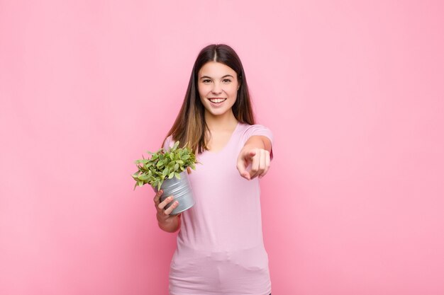 Giovane donna graziosa che indica alla macchina fotografica con un sorriso soddisfatto, sicuro, amichevole, scegliendovi con una pianta