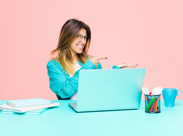 Giovane donna graziosa che lavora con un computer portatile che sorride allegramente e che indica lo spazio della copia sulla palma dal lato che mostra o che annuncia un oggetto