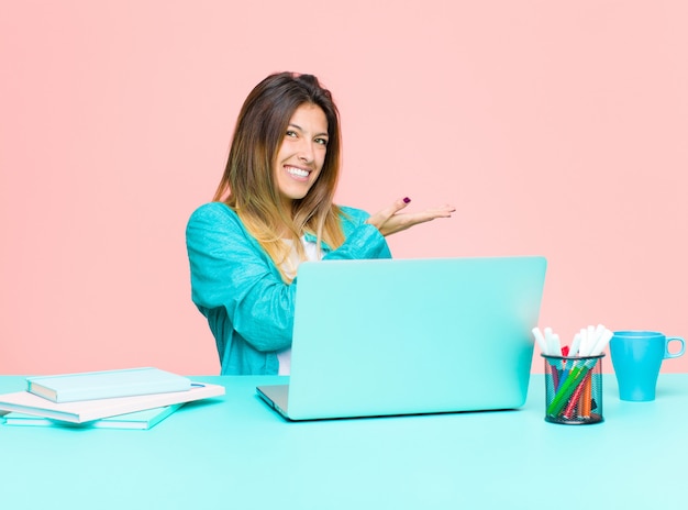 Giovane donna graziosa che lavora con un computer portatile che sorride allegramente, sentendosi felice e mostrando un concetto nello spazio della copia con il palmo della mano