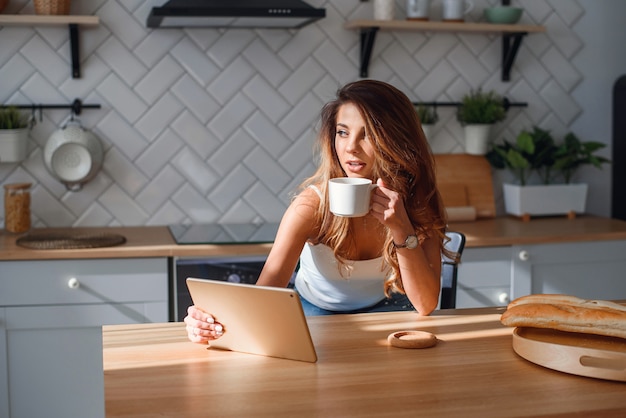 Giovane donna graziosa che per mezzo della compressa mentre bevendo il caffè di mattina dopo la prima colazione sulla cucina.