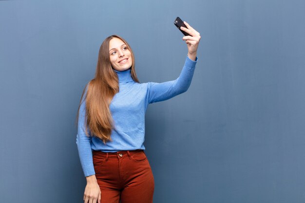 Giovane donna graziosa che per mezzo di uno Smart Phone contro la parete blu con un copyspace