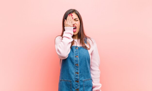 Giovane donna graziosa che sembra assonnata, annoiata e che sbadiglia, con un mal di testa e una mano che copre metà del viso contro il muro rosa