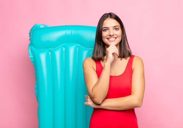 Giovane donna graziosa che sembra felice e sorridente con la mano sul mento, chiedendosi o facendo una domanda, confrontando le opzioni