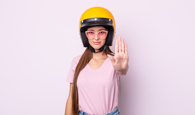 Giovane donna graziosa che sembra seria mostrando palmo aperto che fa gesto di arresto. motociclista e casco