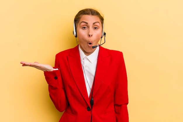 Giovane donna graziosa che sembra sorpresa e scioccata con la mascella caduta in possesso di un concetto di telemarketing di oggetti