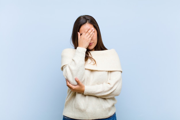 Giovane donna graziosa che sembra stressata, vergogna o turbata, con un mal di testa, che copre il viso con la mano contro il muro blu