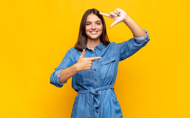 Giovane donna graziosa che si sente felice, amichevole e positiva, sorride e fa un ritratto o una cornice per foto con le mani