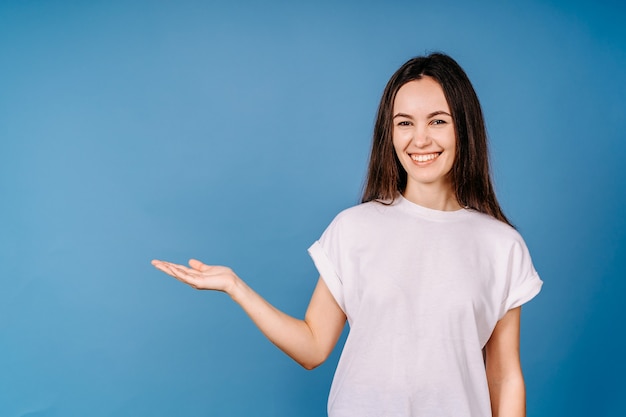 Giovane donna graziosa che si sente felice e sorridente casualmente, guardando un oggetto o un concetto tenuto sulla mano sul lato contro il muro blu