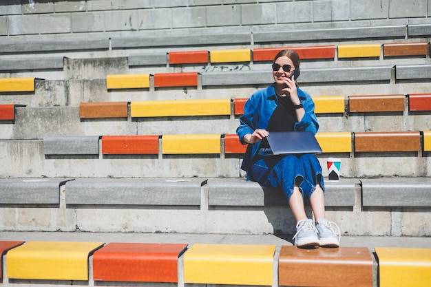 Giovane donna graziosa che si siede con il computer portatile in una strada moderna del parco che utilizza lo smartphone sul lavoro IT remoto in stile estivo casual sorridente con gli occhiali