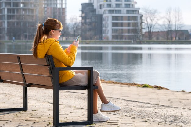 Giovane donna graziosa che si siede su una panchina del parco che sfoglia il suo sellphone all'aperto la sera