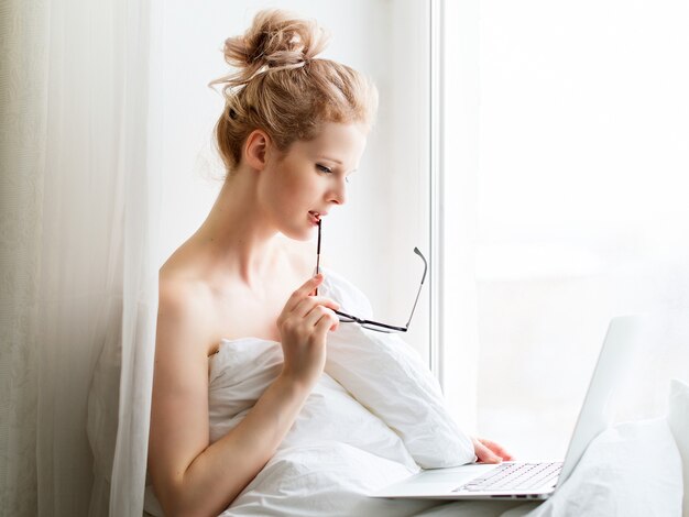 Giovane donna graziosa che si siede sulla finestra con il computer portatile e il lavoro