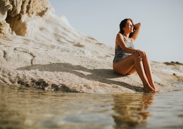 Giovane donna graziosa che si siede sulla riva rocciosa dal mare