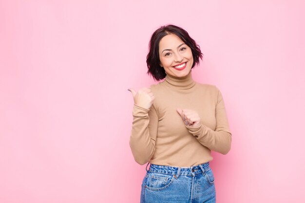 Giovane donna graziosa che sorride allegramente e casualmente che punta a copiare lo spazio sul lato, sentendosi felice e soddisfatto contro il muro rosa