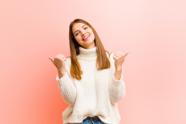 Giovane donna graziosa che sorride con gioia e che sembra felice, sentendosi spensierata e positiva con entrambi i pollici contro la parete rosa