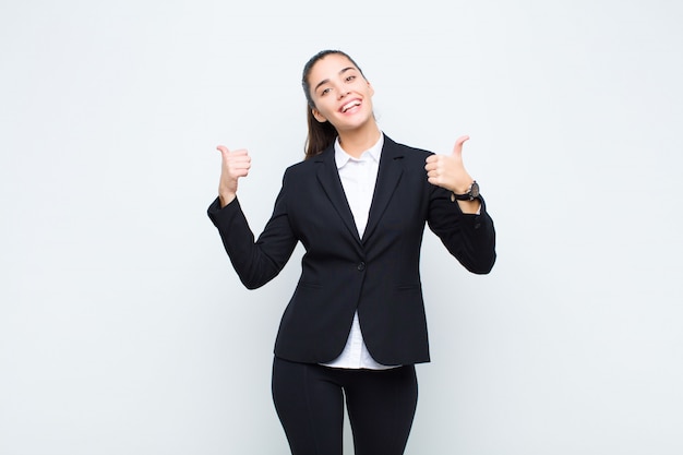 Giovane donna graziosa che sorride con gioia e sembra felice, sentendosi spensierata e positiva con entrambi i pollici in su il concetto di business