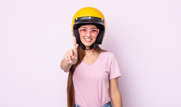 Giovane donna graziosa che sorride con orgoglio e sicurezza facendo il motociclista e il casco numero uno