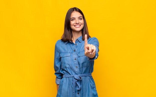 Giovane donna graziosa che sorride con orgoglio e sicurezza facendo posare trionfante il numero uno, sentendosi come un leader