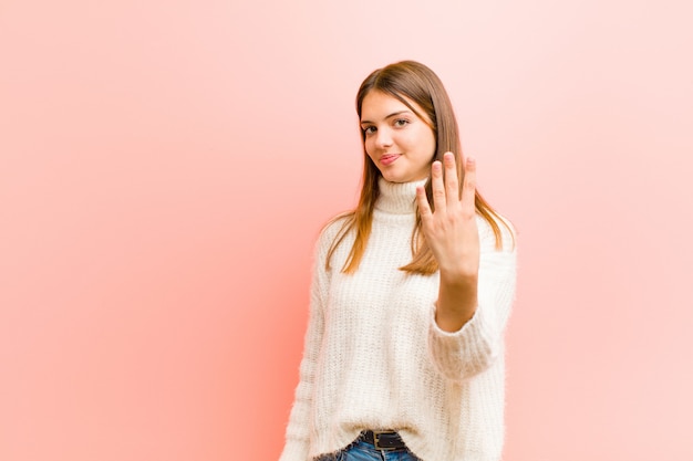 Giovane donna graziosa che sorride e che sembra amichevole, mostrando numero quattro o quarto con la mano in avanti, conto alla rovescia contro la parete rosa