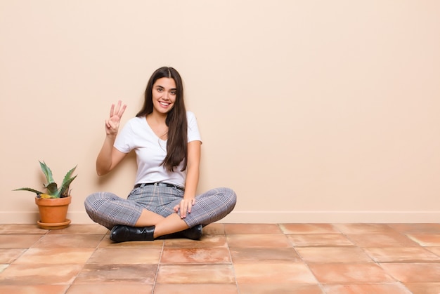 Giovane donna graziosa che sorride e che sembra amichevole, mostrando numero tre o terzo con la mano in avanti, contando alla rovescia seduta su un pavimento