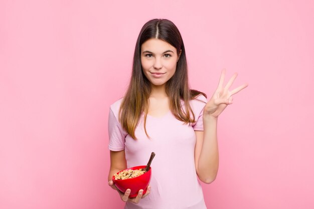 Giovane donna graziosa che sorride e che sembra amichevole, mostrando numero tre o terzo con la mano in avanti, conto alla rovescia con una ciotola della prima colazione