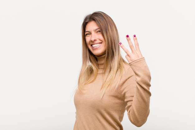 Giovane donna graziosa che sorride e che sembra amichevole, mostrando numero tre o terzo con la mano in avanti, conto alla rovescia contro il fondo bianco