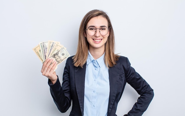 Giovane donna graziosa che sorride felicemente con una mano sull'anca e concetto di banconote in dollari fiducioso