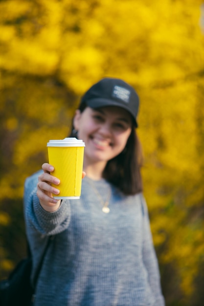 Giovane donna graziosa che tiene e mostra la tazza di carta del caffè gialla