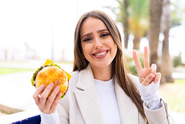 Giovane donna graziosa che tiene un hamburger all'aperto che sorride e che mostra il segno di vittoria