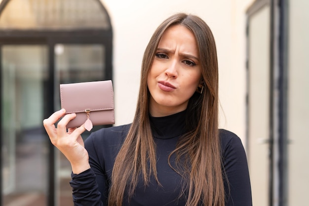 Giovane donna graziosa che tiene un portafoglio all'aperto con espressione triste