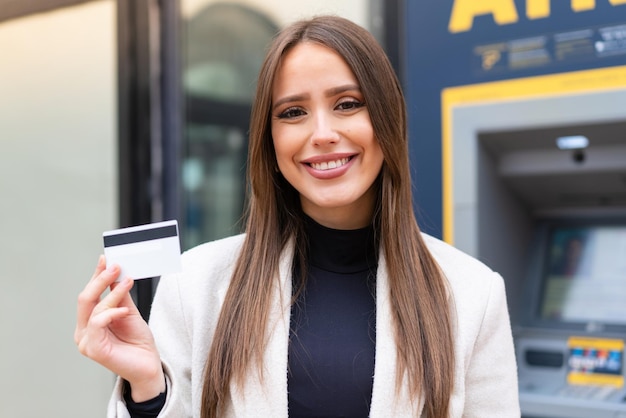 Giovane donna graziosa che tiene una carta di credito all'aperto che sorride molto