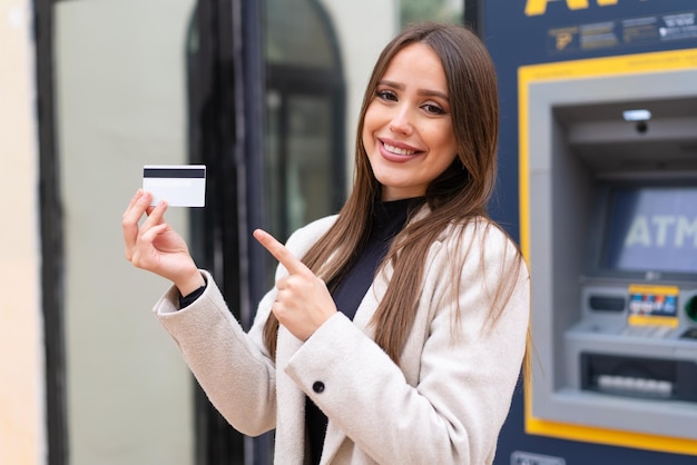 Giovane donna graziosa che tiene una carta di credito all'aperto e che la indica