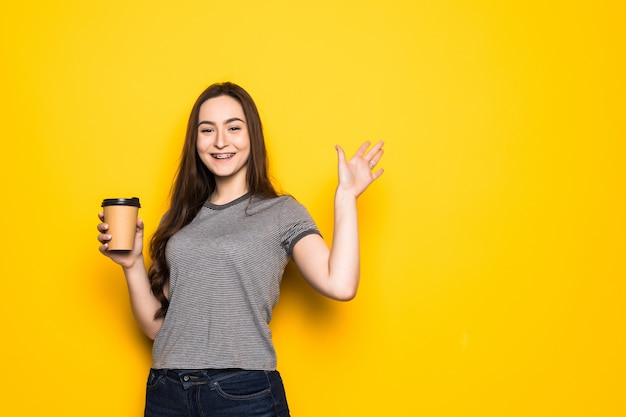 Giovane donna graziosa con la tazza di caffè che fluttua con le mani sulla parete gialla