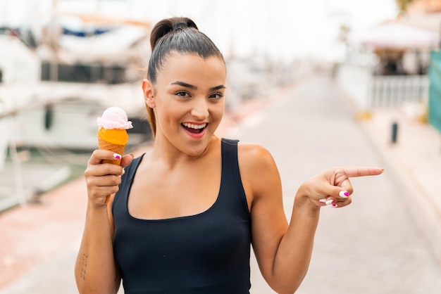 Giovane donna graziosa con un gelato alla cornetta all'aperto sorpresa e che punta il dito verso il lato