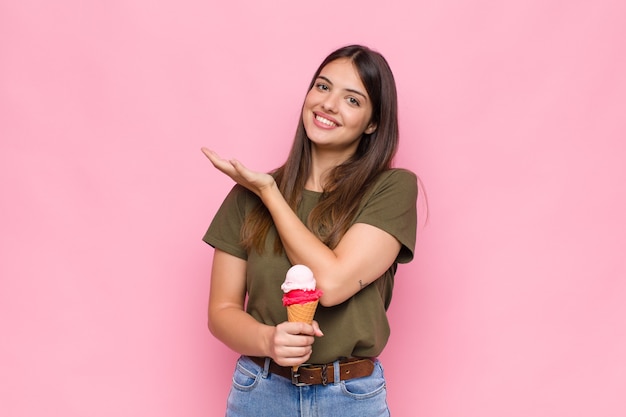 Giovane donna graziosa con un gelato che sorride allegramente, sentendosi felice e mostrando un concetto nello spazio della copia con il palmo della mano