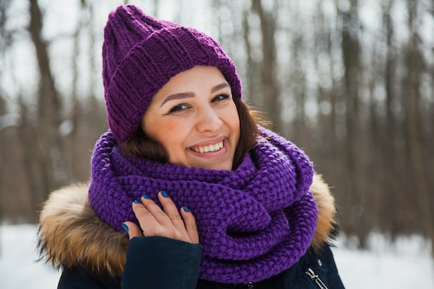 Giovane donna graziosa con un sorriso felice in snood viola e cappello lavorato a maglia. Giornata invernale nevosa, tempo gelido.